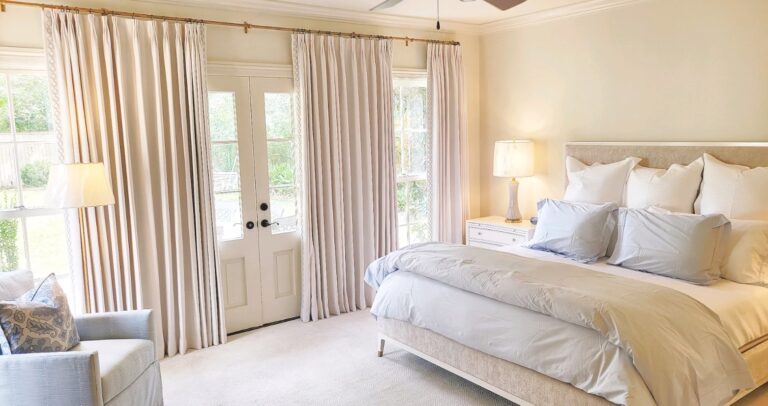 A bedroom with a large bed, light blue bedding, a padded headboard, two table lamps, an armchair, and curtains covering glass doors and windows.
