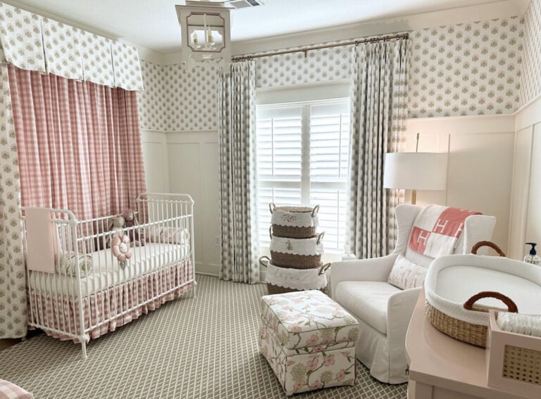 Nursery with a white crib, pink and white bedding, floral armchair, and a window with patterned curtains. Changing table is beside an armchair.