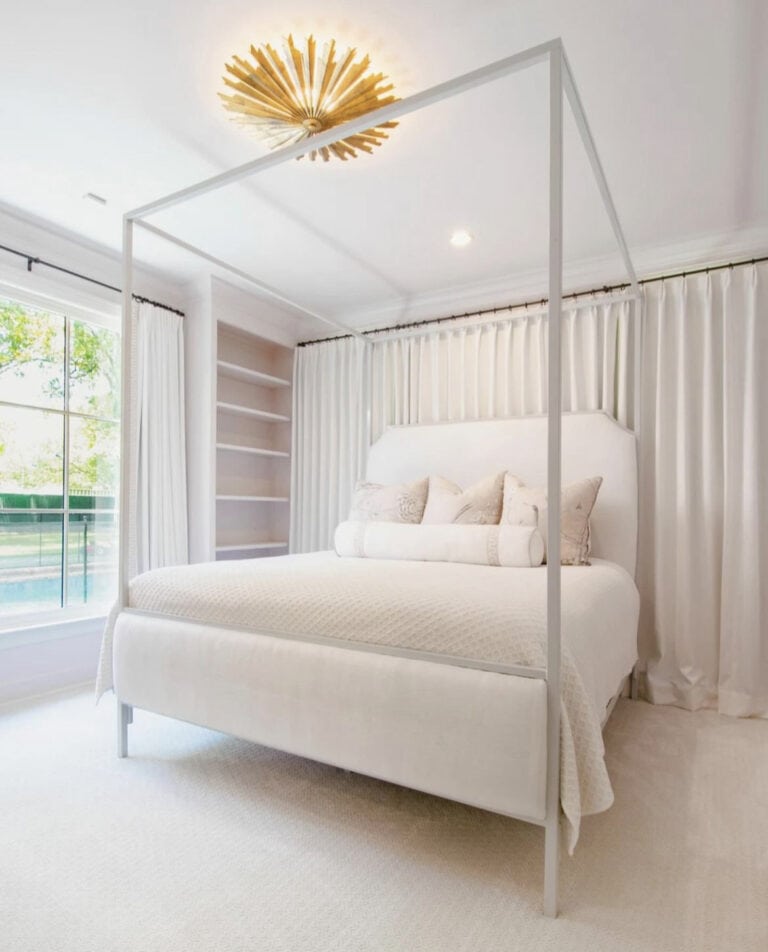 A bedroom with a white canopy bed, white bedding, curtains, built-in shelves, and a sunburst chandelier. A large window is on the left.