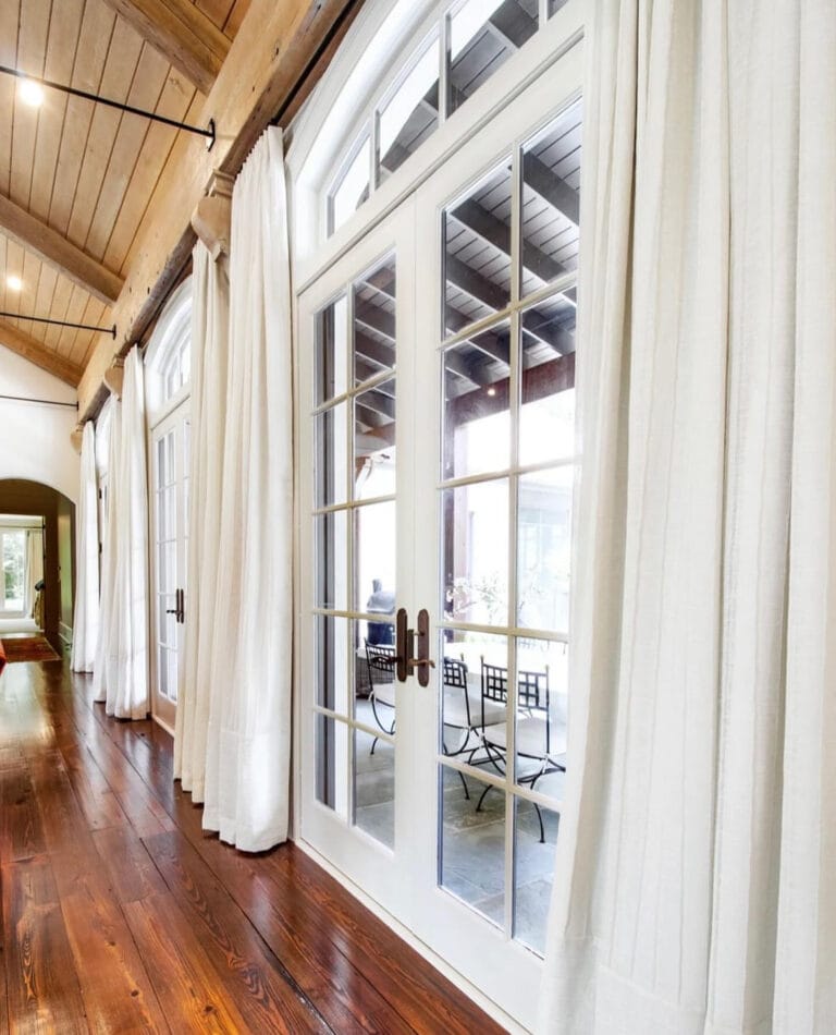 A row of large, arched French doors with white drapes open to a patio with metal chairs. The room features wooden floors and a vaulted ceiling.