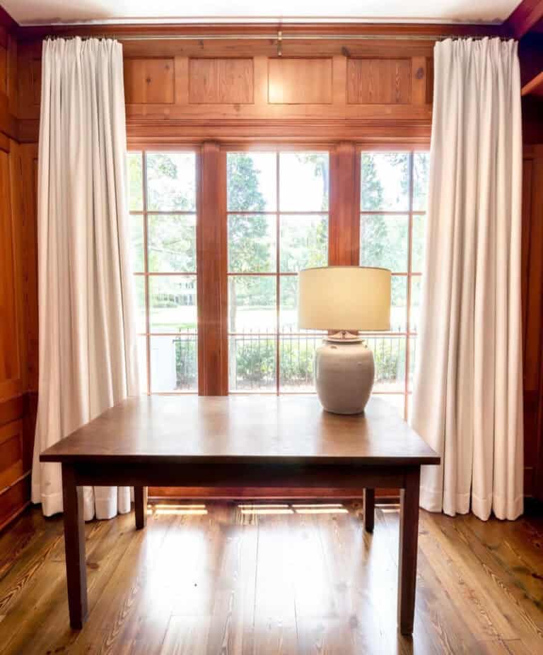 A wooden table with a beige lamp sits in front of tall windows with white curtains in a wood-paneled room. Sunlight streams through the glass.