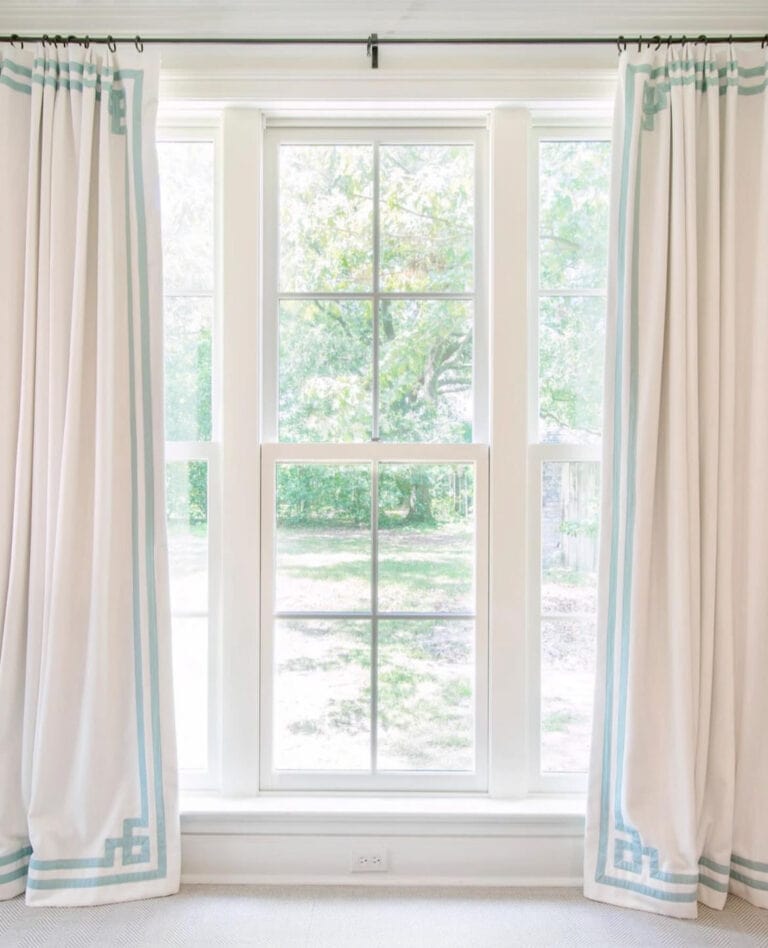 Large window with six panes, framed by white curtains with blue geometric trim, revealing a view of green trees and grass outside.