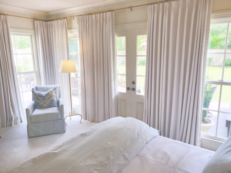 A bright bedroom with white curtains, an armchair with a patterned cushion, a standing lamp, and a bed. Glass doors lead to a garden.