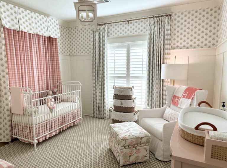 A nursery with a crib, armchair, and changing table. Decor features pink and white patterns, floral curtains, and a basket with stuffed animals.