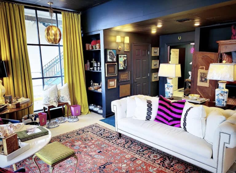 Living room with a white sofa, colorful pillows, a patterned rug, paintings, lamps, and mustard curtains. Shelves with decor items line the walls.