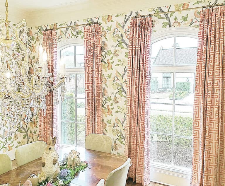 Dining room with floral wallpaper, red-patterned curtains, arched windows, crystal chandelier, and a wooden table with decorative items.