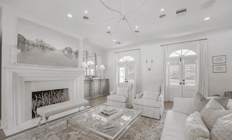 Elegant living room with white furniture, a glass coffee table, and a decorative fireplace. Large painting hangs above the fireplace.