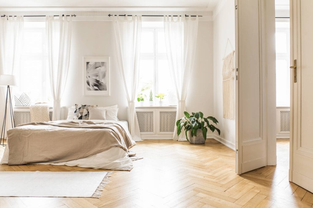 A bright bedroom with a neatly made bed, white curtains, framed artwork, a potted plant, and parquet flooring, lit by sunlight through large windows.