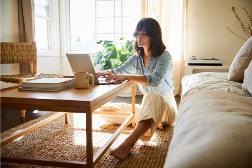 Woman sitting on the floor in a sunlit room, typing on a laptop on a wooden coffee table, with books and a mug nearby.